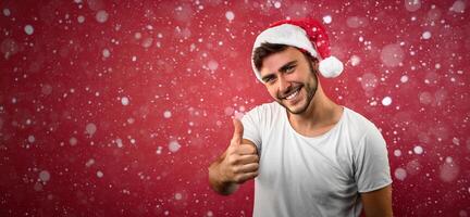 Young handsome caucasian guy in a white t-shirt and Santa hats stands on red background in studio and showing thumbs up. photo