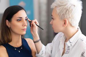 Make-up artist applying bright base color eyeshadow on model's eye and holding a shell with eyeshadow on background photo