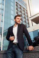 handsome young businessman with a beard and in a business suit standing on the street against the background of the office building next to a comfortable stylish leather bag. photo