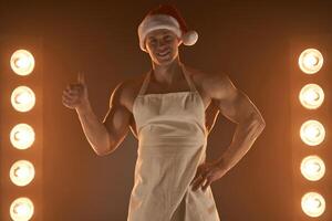 caliente Navidad postre. muscular hombre vistiendo delantal y Papa Noel sombrero demostración pulgar arriba gesto y sonriente a cámara, lamparas iluminación en antecedentes foto