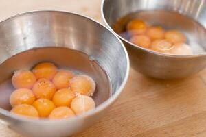 Two stainless steel bowls filled with many cracked eggs with whole yolks, in the wooden table. Close up photo