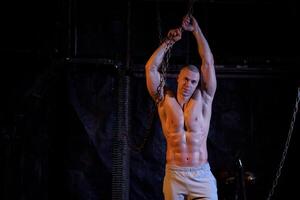 Young shirtless muscular man standing among metal chains, looking at camera, copy space photo