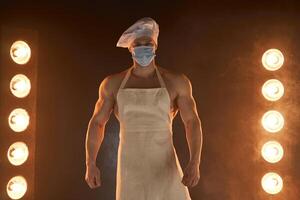 nuevo normal concepto. muscular cocinero vistiendo protector médico máscara delantal y cocinero sombrero, en pie en ahumado antecedentes y lámpara iluminación foto