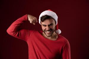 Young handsome caucasian guy in a red sweater and Santa hats stands on red background in studio and showing biceps power photo