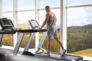 Muscular athletic bodybuilder fitness model running treadmill gym near big window photo