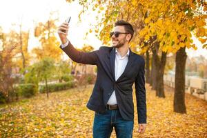 hermoso joven de moda hombre en un elegante negocio traje y Gafas de sol hacer selfie en su teléfono caminando en el otoño parque en el antecedentes de brillante amarillo hojas. foto