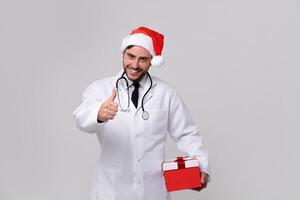 Young handsome doctor in white uniforme and Santa Claus hat standing in studio on red background smile and finger in camera photo