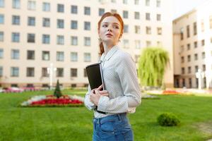 un hermosa Pelirrojo niña con pecas sostiene un cuaderno en su manos y va a colega. preparación para exámenes espalda a escuela. retrato de un estudiante foto