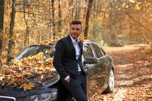 un hermoso joven caucásico hombre vistiendo negocio traje en pie cerca negro coche en naturaleza. foto