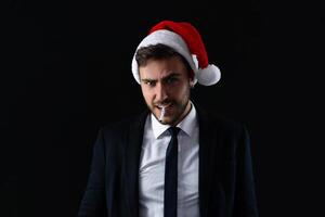 Young handsome caucasian guy in business suit and Santa hats stands on black background in studio with serious face and smoking cigarette photo