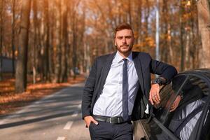 A handsome young Caucasian man wearing business suit standing near black car on nature. photo