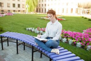 Young beautiful red-haired girl with freckles sitting on a bench near the university writing in a notebook with homework. Portrait of a student. Back to school concept. photo