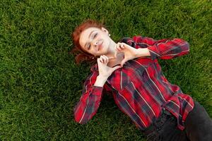 Beautiful red-haired young student with freckles lying on her back on the grass top view. Dressed in a red checked shirt, reveals a heart of fingers Love and positive photo