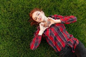 hermosa Pelirrojo joven estudiante con pecas acostado en su espalda en el césped parte superior vista. vestido en un rojo comprobado camisa, revela un corazón de dedos amor y positivo foto