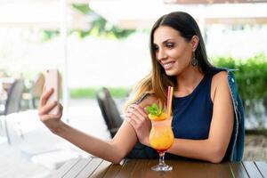 A beautiful smiling woman is drinking an exotic cocktail in an outdoor restaurant and is holding a mobile phone. photo