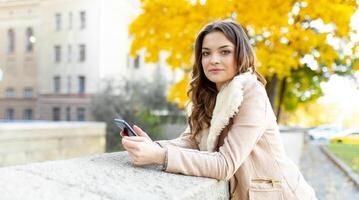 Beautiful caucasian brunette girl standing warm autumn day with background of trees with yellow foliage and a city. Dressed wool sweater and jacket. photo