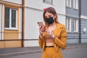 Young beautiful caucasian girl 20 years wear black face mask protection against epidemic coronavirus covid-19 walks down the street looks at the phone screen. photo