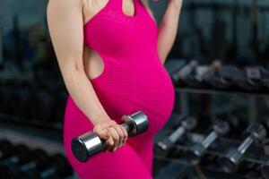 Pregnant woman lifting up dumbbells training biceps muscle at the gym standing near mirror photo