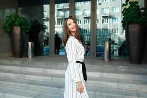 alegre bonito joven dama entrando vaso puerta dentro moderno oficina, hotel, cafetería, negocio centrar y mirando espalda a ola adiós. niña vistiendo elegante anteojos, y sonriente espalda foto