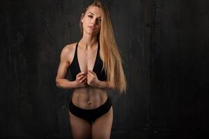 Image of muscular young female athlete wearing black sport wear standing looking in camera on dark background. Woman bodybuilder resting after workout. photo