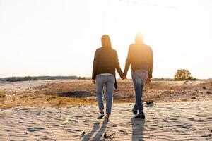 Young European couple dressed leather jacket and blue jeans walking desert holding hands view from the back. photo