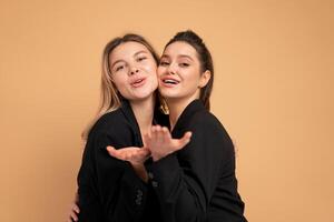 Two young business woman dressed black suit laugh and happy. Two beautiful caucasian female business person hugging isolated on beige color background photo