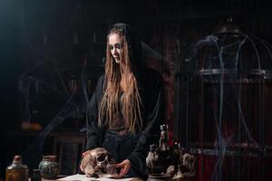 Halloween concept. Witch portrait close up with dreadlocks dressed black hood standing dark room with cage on background photo