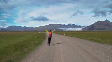 weiblich Tourist mit Rucksack geht durch Schmutz Straße. sie ist glücklich und heiter. schön Aussicht von das Berge und Gletscher von Island. Freiheit und Reise Konzept. 4k video