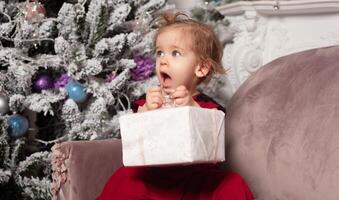 A beautiful cute little girl dressed in an elegant evening red dress sits on the couch and opens a New Year's gift. With Christmas tree on the background photo