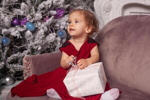 un hermosa linda pequeño niña vestido en un elegante noche rojo vestir se sienta en el sofá y abre un nuevo años regalo. con Navidad árbol en el antecedentes foto