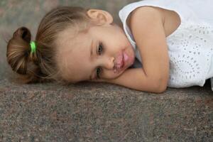 A beautiful little girl of European appearance blonde hair lying sad on street asphalt. photo