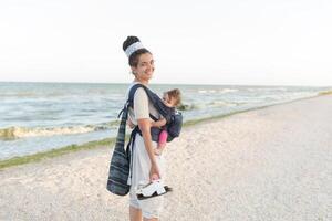 un pequeño niño se sienta en un mochila y camina a lo largo con el madre a lo largo el costa. verano familia vacaciones concepto foto