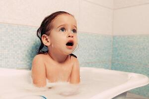 divertido alegre contento niñito bebé tomando un bañera jugando con espuma burbujas pequeño niño en un bañera. sonriente niño en baño en azul antecedentes. higiene y salud cuidado. foto