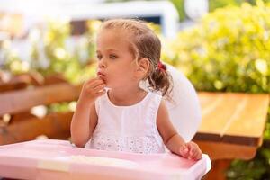 linda pequeño caucásico niña comiendo espaguetis a mesa sentado en niño asiento al aire libre restaurante. foto