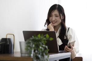 un japonés mujer comprobación teléfono inteligente por remoto trabajo en el pequeño oficina foto