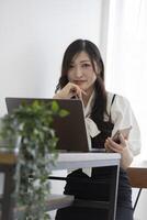 un japonés mujer comprobación teléfono inteligente por remoto trabajo en el pequeño oficina foto