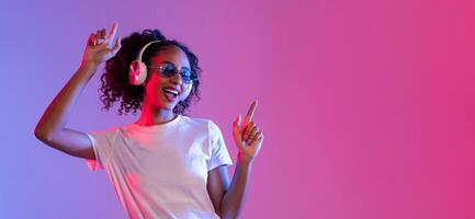 Joyful black woman with sunglasses and headphones dancing on neon background, free space photo