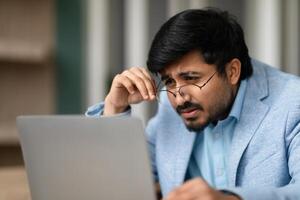 Indian businessman squinting through eyeglasses looking at laptop in office photo