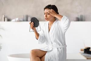 Young black woman in white bathrobe holding mirror, critically examining her skin photo