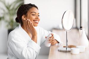 Smiling young black woman in white bathrobe applying skincare product photo