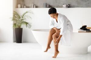 Smiling black woman in bathrobe dry brushing legs, practicing skin exfoliation routine photo