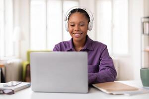 sonriente hembra negro estudiante en auriculares utilizando ordenador portátil a escritorio a hogar foto