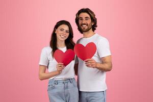 Happy couple in white t-shirts and jeans holding red heart-shaped paper cutouts photo