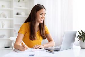 sonriente asiático mujer trabajando en su ordenador portátil y tomando notas a hogar foto