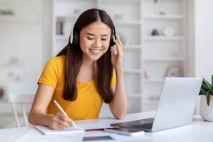 Smiling, young asian woman wearing headphones taking notes and using her laptop photo