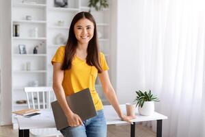 Smiling young asian woman holding closed laptop, standing in well-lit home office photo