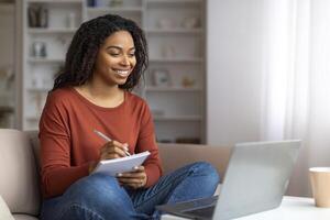 Smiling young black woman writing down notes from her laptop session photo