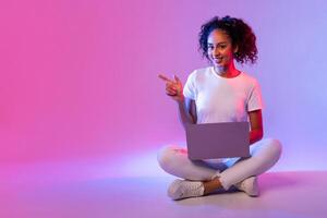 Smiling black lady with laptop sitting cross-legged, pointing to side at free space photo