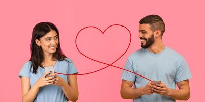 A young woman with a gentle smile, holding a smartphone, glances at a bearded man also with a phone photo