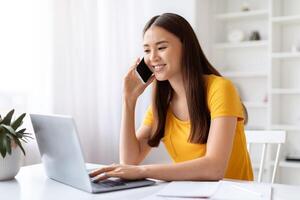 Happy asian woman talking on cellphone while working on laptop at home photo